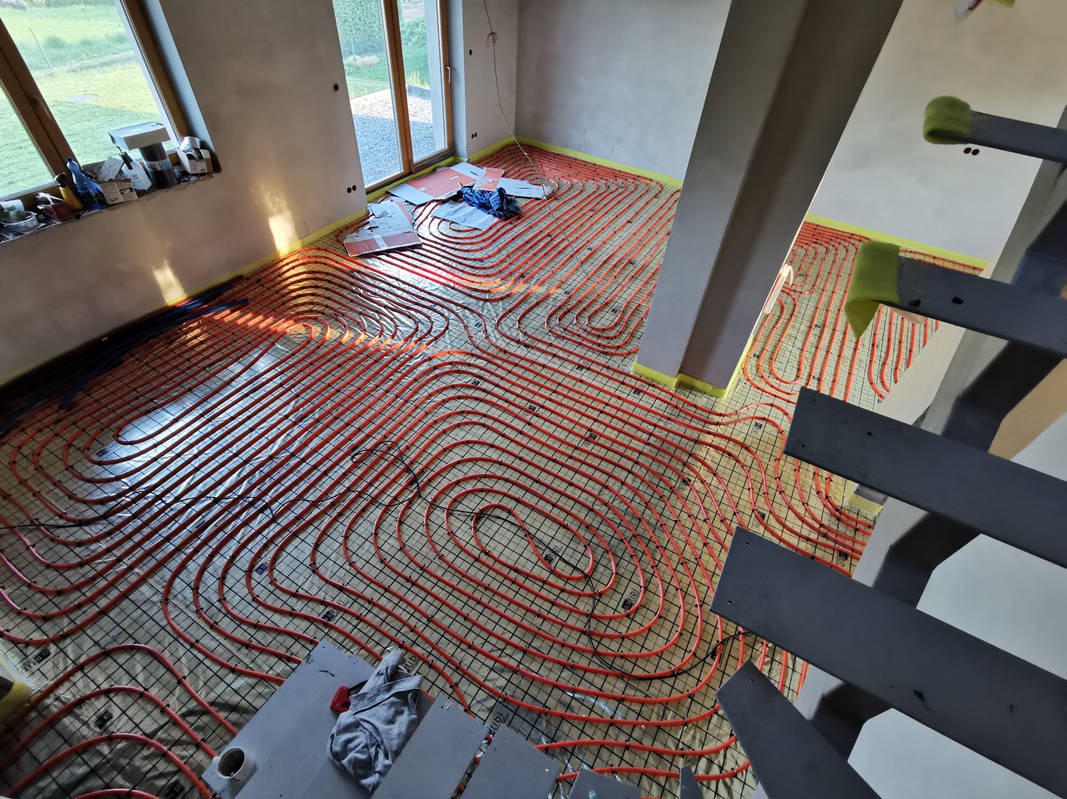Instalacja ogrzewania podłogowego: czerwone rury ułożone na siatce w jasnym pomieszczeniu z dużymi oknami, widok z góry na etap prac przed wylaniem posadzki.