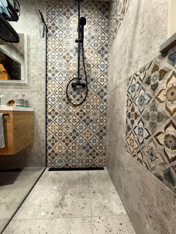 Wykończona kabina prysznicowa z ozdobnymi, wzorzystymi płytkami na ścianach i podłodze. Widoczna armatura w kolorze czarnym. Kabina bez brodzika z odpływem liniowym.