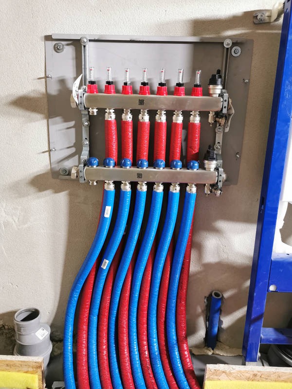 Rozdzielacz ogrzewania podłogowego z czerwono-niebieskimi rurami w surowym wnętrzu. Widoczne elementy montażowe i przyłącza. Instalacja w trakcie realizacji.
