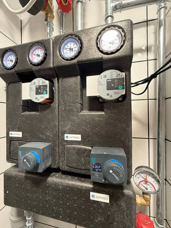 Dwa czarne moduły AFRISO z manometrami i regulatorami temperatury, część instalacji grzewczej w pomieszczeniu z białymi płytkami. Widoczne rury i zawory.