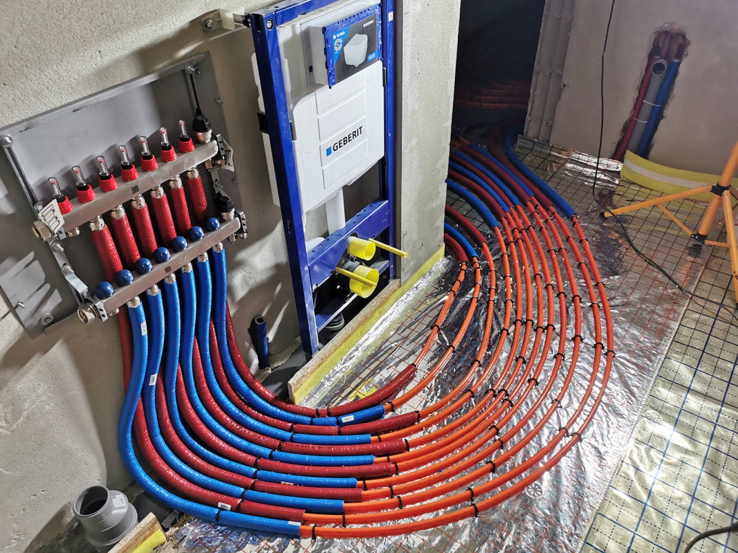 Rozdzielacz ogrzewania podłogowego z rurami w kolorze czerwonym i niebieskim, ułożonymi na srebrnej folii izolacyjnej, obok stelaża Geberit. Widok z góry, w trakcie montażu.