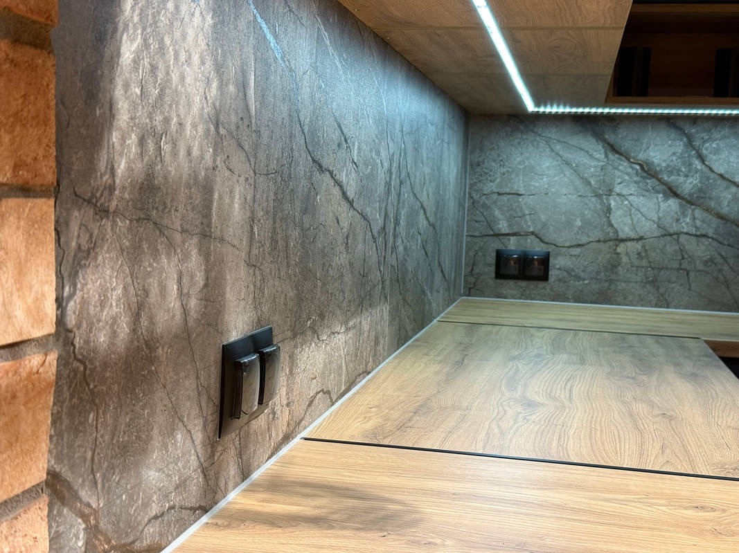 Wykończenie ściany imitujące kamień z widocznymi gniazdkami elektrycznymi i oświetleniem LED podszafkowym w kuchni. Blat drewnopodobny.