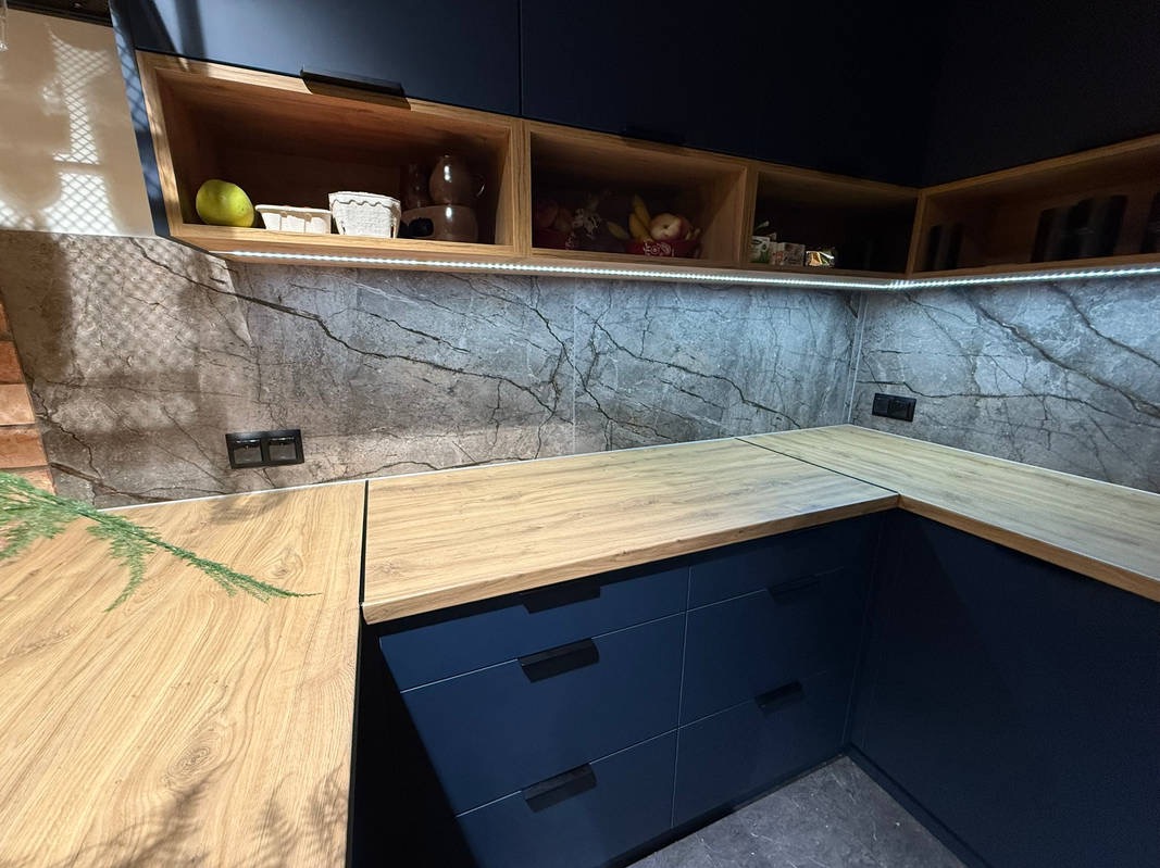 Nowoczesna kuchnia z ciemnymi szafkami, drewnianym blatem i marmurowymi płytkami. Oświetlenie LED podkreśla wzór płytek. Widoczne gniazdka elektryczne.