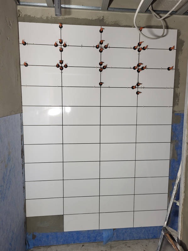 Układanie białych, błyszczących płytek na ścianie z użyciem poziomych i pionowych krzyżyków dystansowych. Widoczne fragmenty drabiny i sufitu.