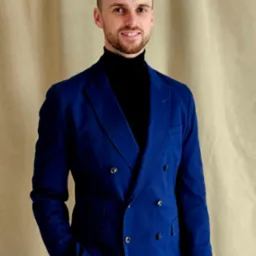 Uśmiechnięty mężczyzna w niebieskiej dwurzędowej marynarce i czarnym golfie, stojący na tle beżowej tkaniny, z dłonią w kieszeni.