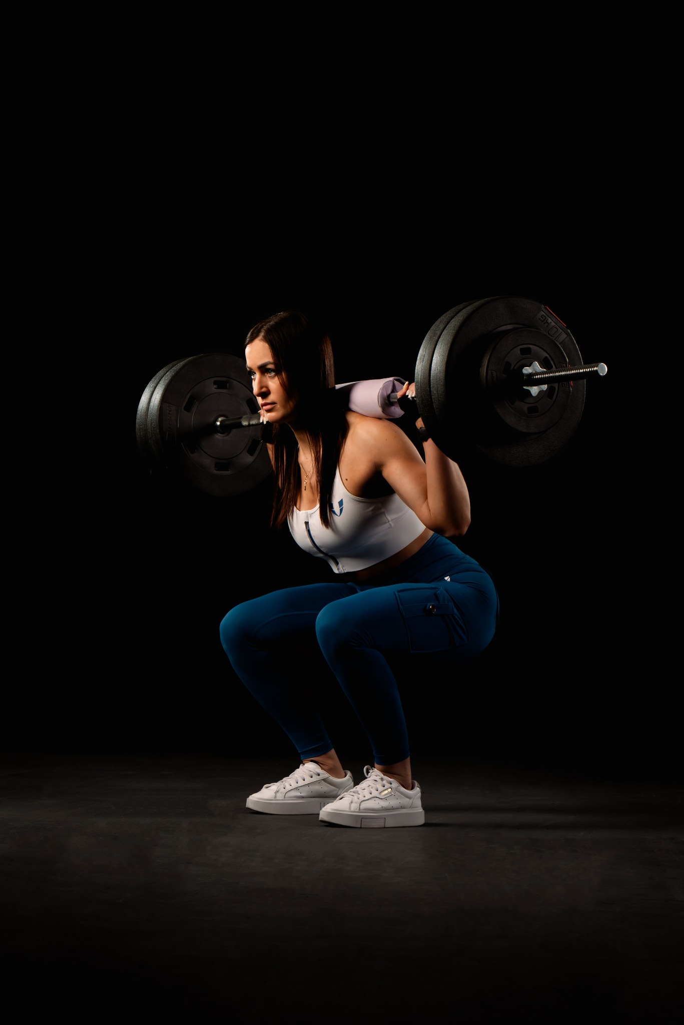 Kobieta w sportowym stroju wykonuje przysiad ze sztangą na plecach w siłowni na czarnym tle, widok z boku.