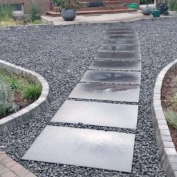 BRUK-GARDEN Leszek Leśniczak - Ścieżka z prostokątnych płyt prowadząca przez szary żwir, obrzeża z kostki betonowej oddzielają żwir od rabat z roślinami ozdobnymi.