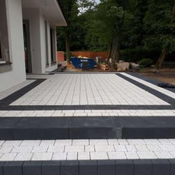 BRUK-GARDEN Leszek Leśniczak - Ułożona nawierzchnia z jasnoszarej kostki brukowej obramowana ciemnoszarą kostką, tworząca schody przed budynkiem. W tle widoczny kontener budowlany i zieleń.