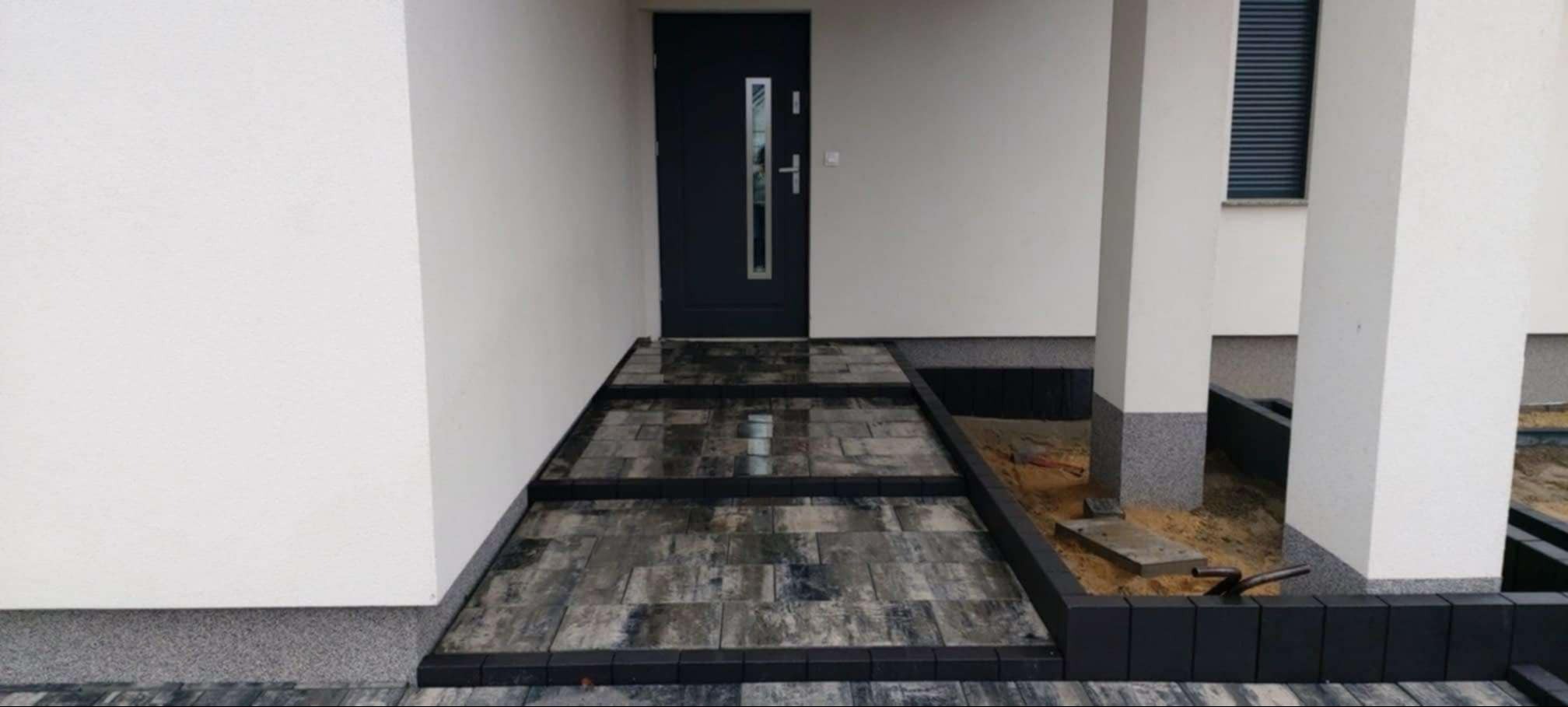 Ułożona ścieżka z kostki brukowej prowadząca do drzwi wejściowych domu, z widocznymi schodkami i obrzeżami z ciemnej kostki. Część terenu obok ścieżki przygotowana pod nasadzenia.