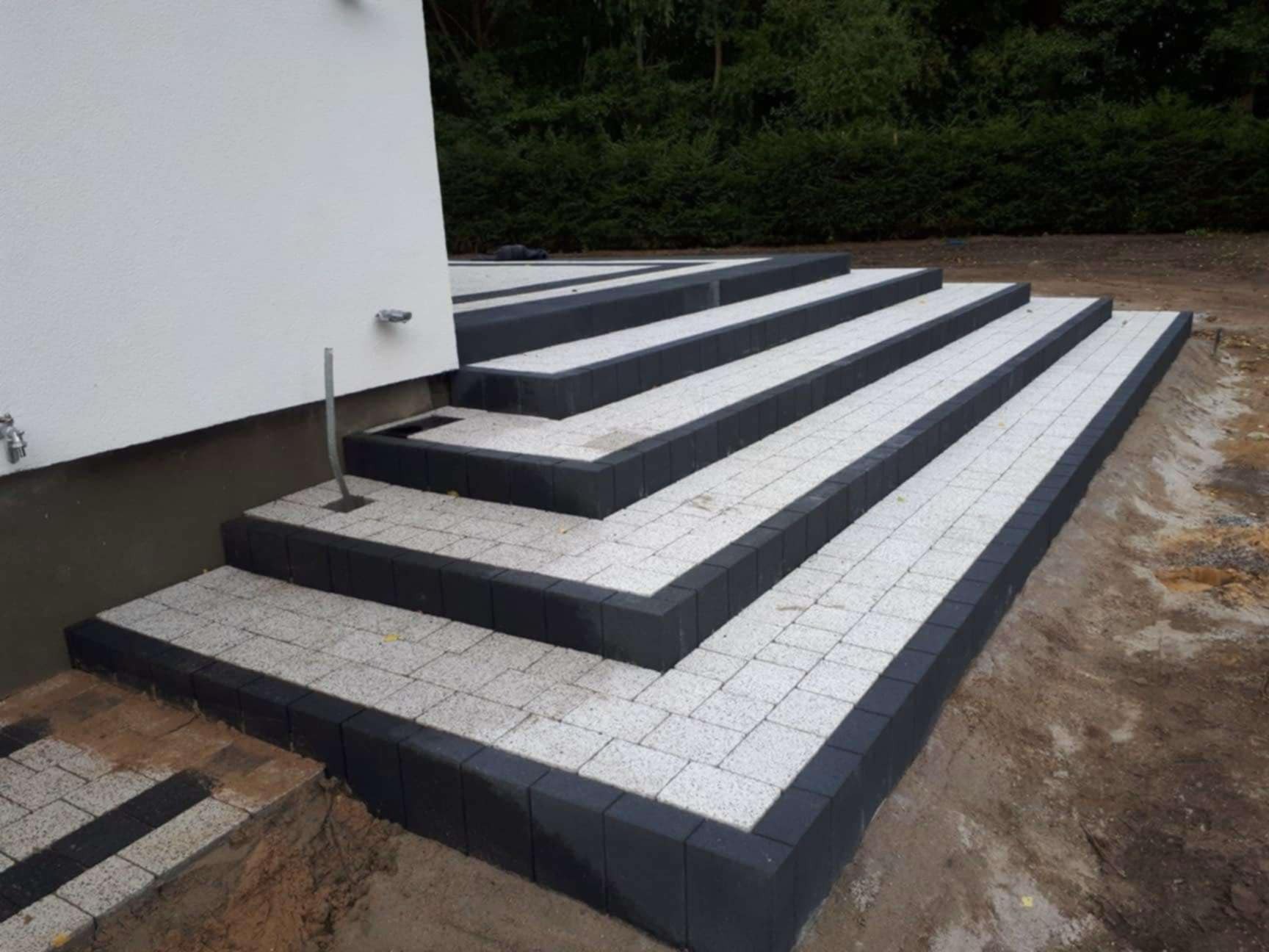 Schody wejściowe wyłożone jasną kostką brukową z ciemnymi obrzeżami, widoczne przyłącza wodne na ścianie budynku