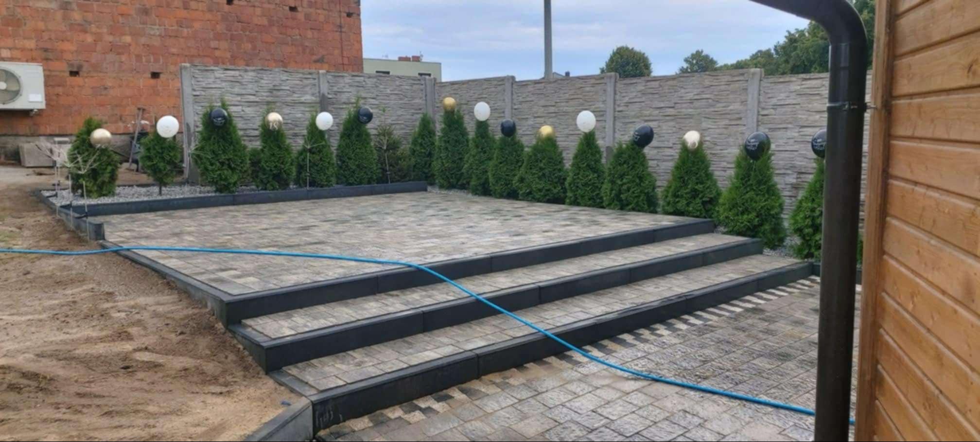 Ułożona kostka brukowa na tarasie z trzema stopniami, obrzeża z ciemnego kamienia, w tle żywopłot z tujek z dekoracjami, widoczny fragment drewnianej ściany i rury spustowej.
