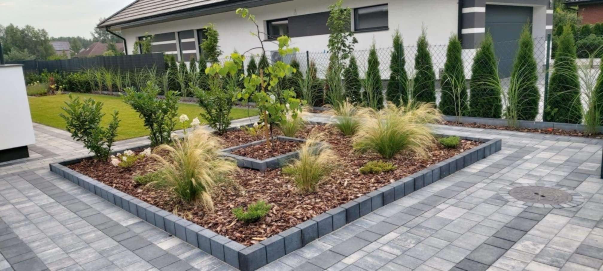 Nowoczesny ogród z rabatą obramowaną kostką, wypełnioną korą, ozdobnymi trawami i młodymi drzewkami, na tle domu i ogrodzenia.
