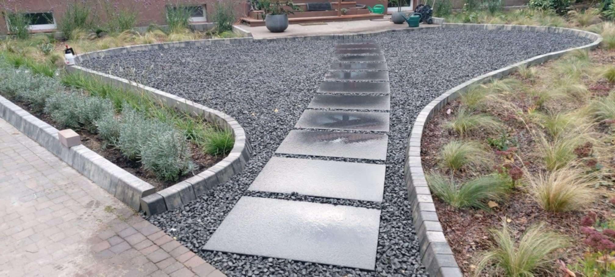 Ścieżka z prostokątnych płyt prowadząca przez szary żwir, obrzeża z kostki betonowej oddzielają żwir od rabat z roślinami ozdobnymi.