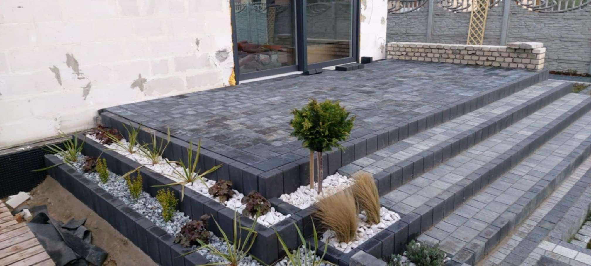 Schody i taras wyłożone szarą kostką brukową, z rabatami kwiatowymi wypełnionymi białym grysem i zielenią, prowadzące do przeszklonych drzwi.