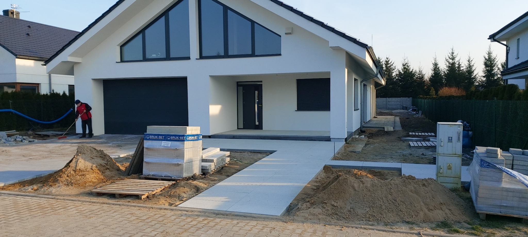 Nowoczesny dom jednorodzinny z jasną elewacją i ciemną stolarką okienną; w trakcie prac wykończeniowych na posesji; widoczne ułożone fragmenty ścieżki z jasnych płyt, paleta z kostką brukową...