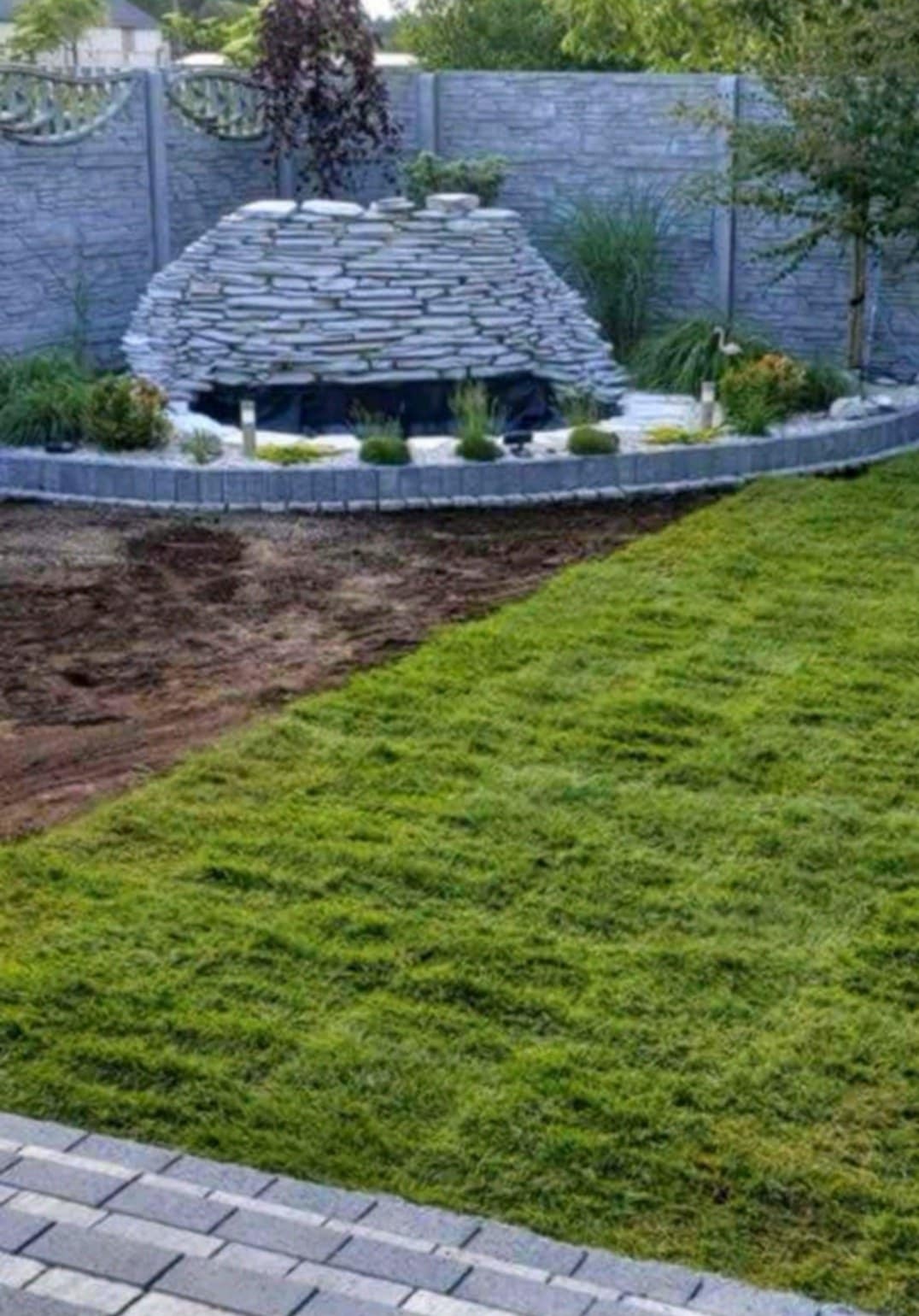 Ogród z elementami dekoracyjnymi: fontanna obłożona kamieniami, rabata z obrzeżem z kostki, trawnik i fragment przygotowanego terenu pod nasadzenia.