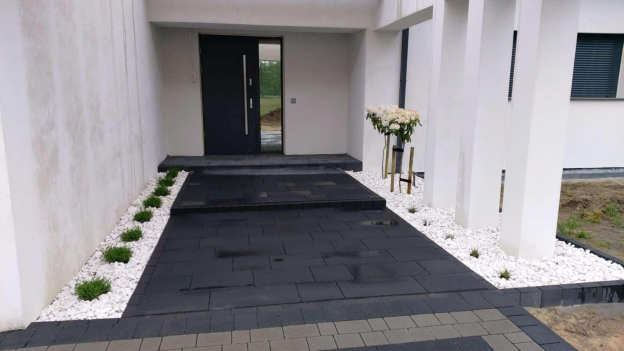 Nowoczesne podejście do domu z ciemnej kostki, obłożone białym grysem i niskimi krzewami, z widoczną elewacją budynku z białymi kolumnami.