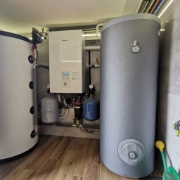 Tomasz Kowalik - Instalacja pompy ciepła Panasonic z dwoma zbiornikami, jeden biały, drugi szary. Widoczne rury, zawory i manometry. Na podłodze panele imitujące drewno.