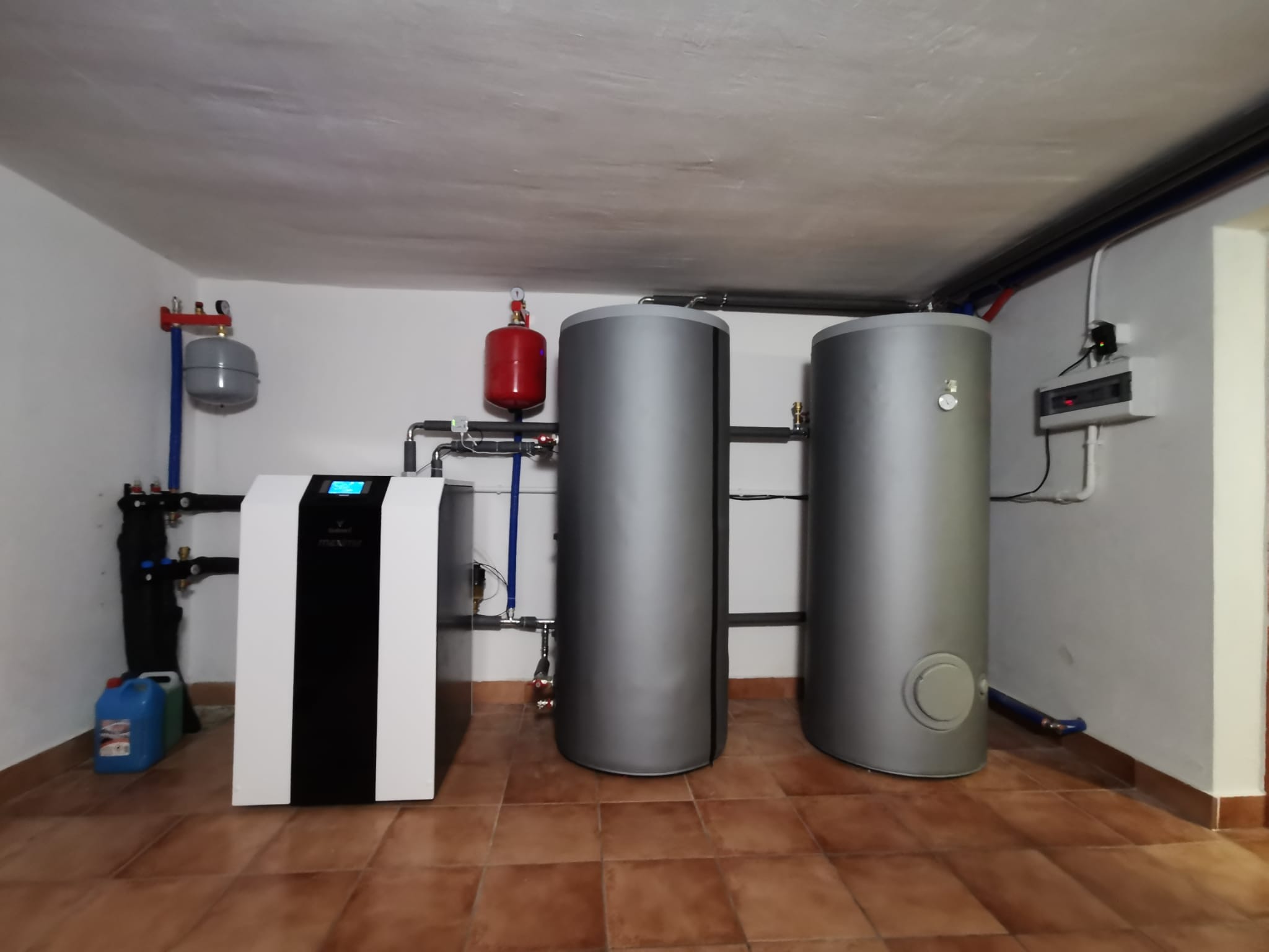 Kotłownia z nowoczesnym piecem dwufunkcyjnym, dwoma szarymi zasobnikami ciepłej wody i instalacją hydrauliczną na brązowych płytkach podłogowych.