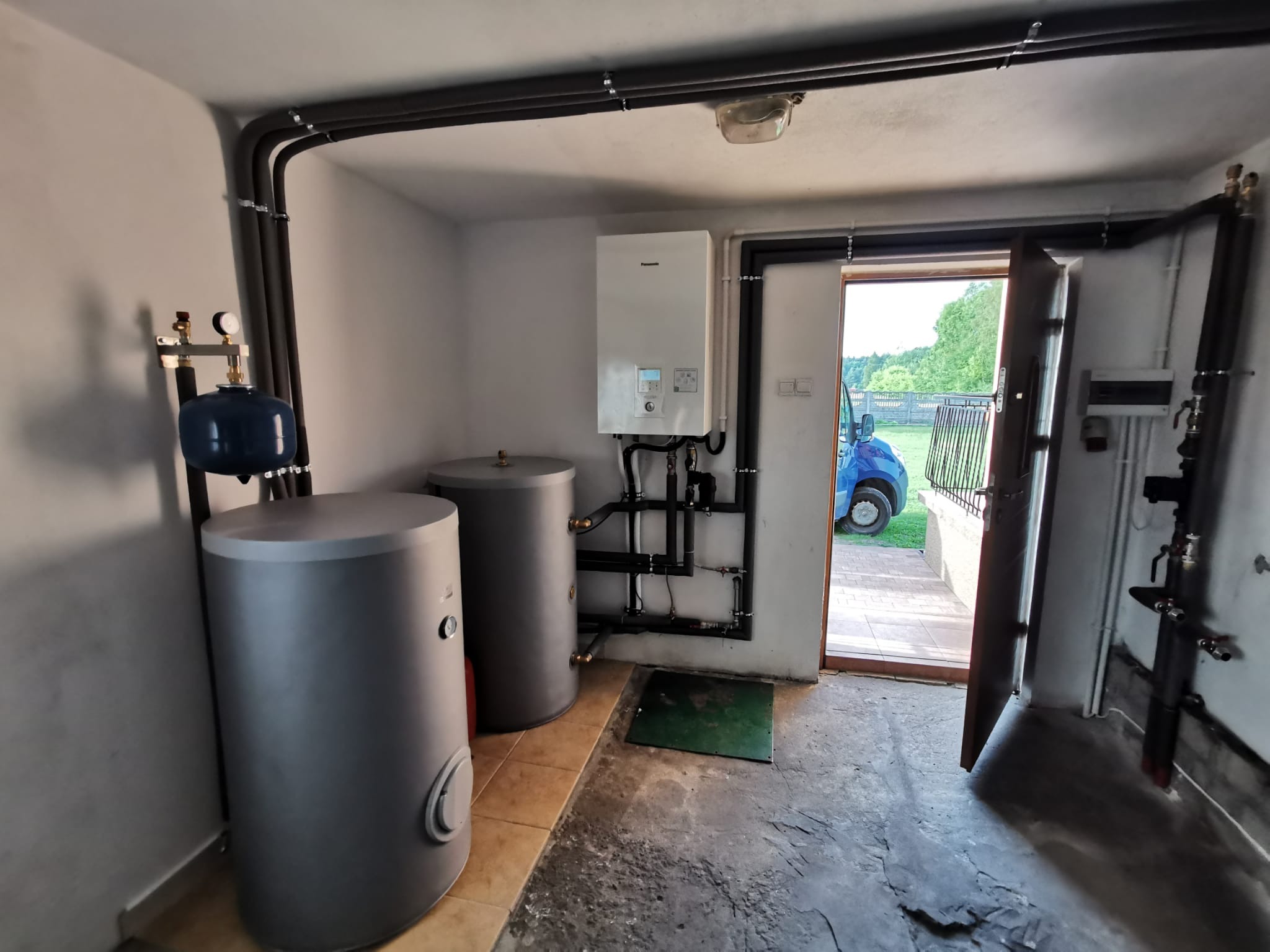 Wnętrze kotłowni z dwoma szarymi zbiornikami, białym piecem gazowym na ścianie i rurami biegnącymi pod sufitem. Otwarta brama garażowa z widokiem na zewnątrz.