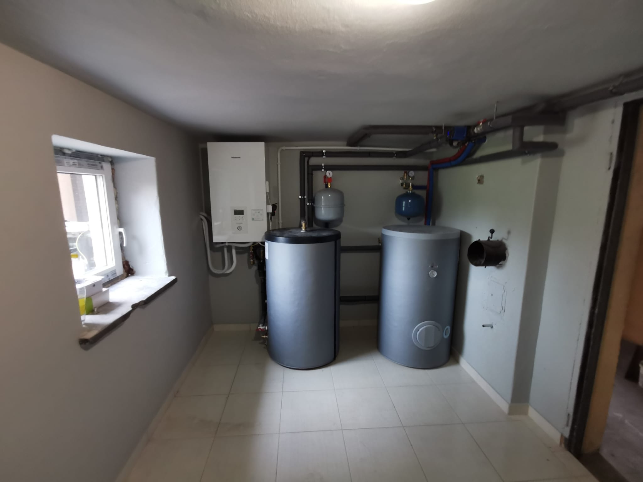 Wnętrze kotłowni z dwoma szarymi zbiornikami, białym piecem gazowym i instalacją rur pod sufitem. Okno po lewej stronie. Jasne płytki na podłodze.