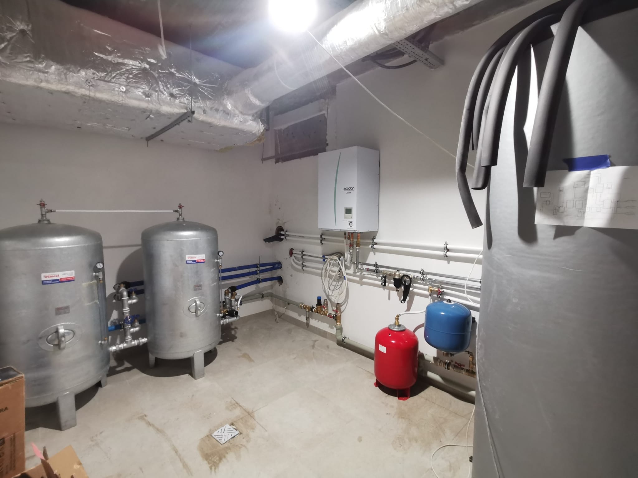 Kompletna instalacja w kotłowni: dwa srebrne zbiorniki, biały piec Ecodan, czerwony i niebieski zbiornik wyrównawczy, rury w piwnicy z surowym wykończeniem.