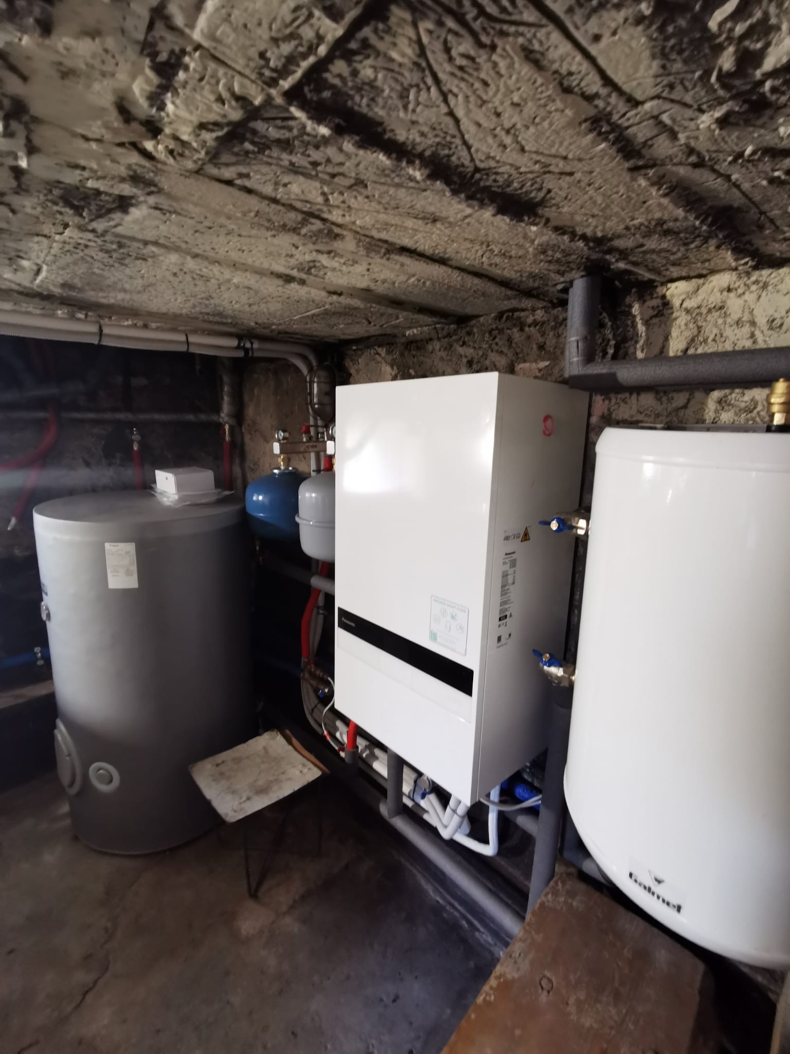 Instalacja pompy ciepła Panasonic w piwnicy: zbiornik buforowy, zasobnik CWU Galmet, biała jednostka wewnętrzna. Widoczne rury i armatura. Surowy strop piwniczny.