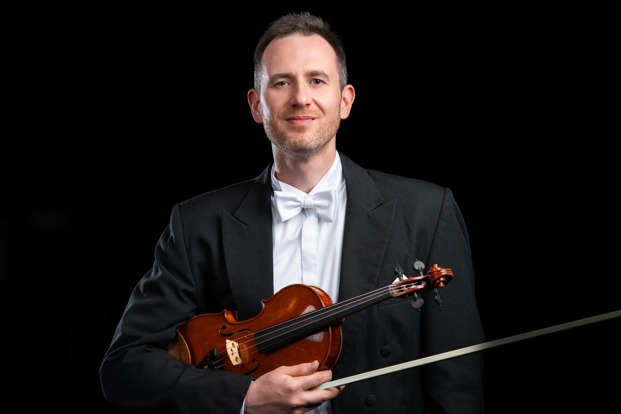 Portret mężczyzny w smokingu z białą muszką, trzymającego skrzypce i smyczek na czarnym tle.