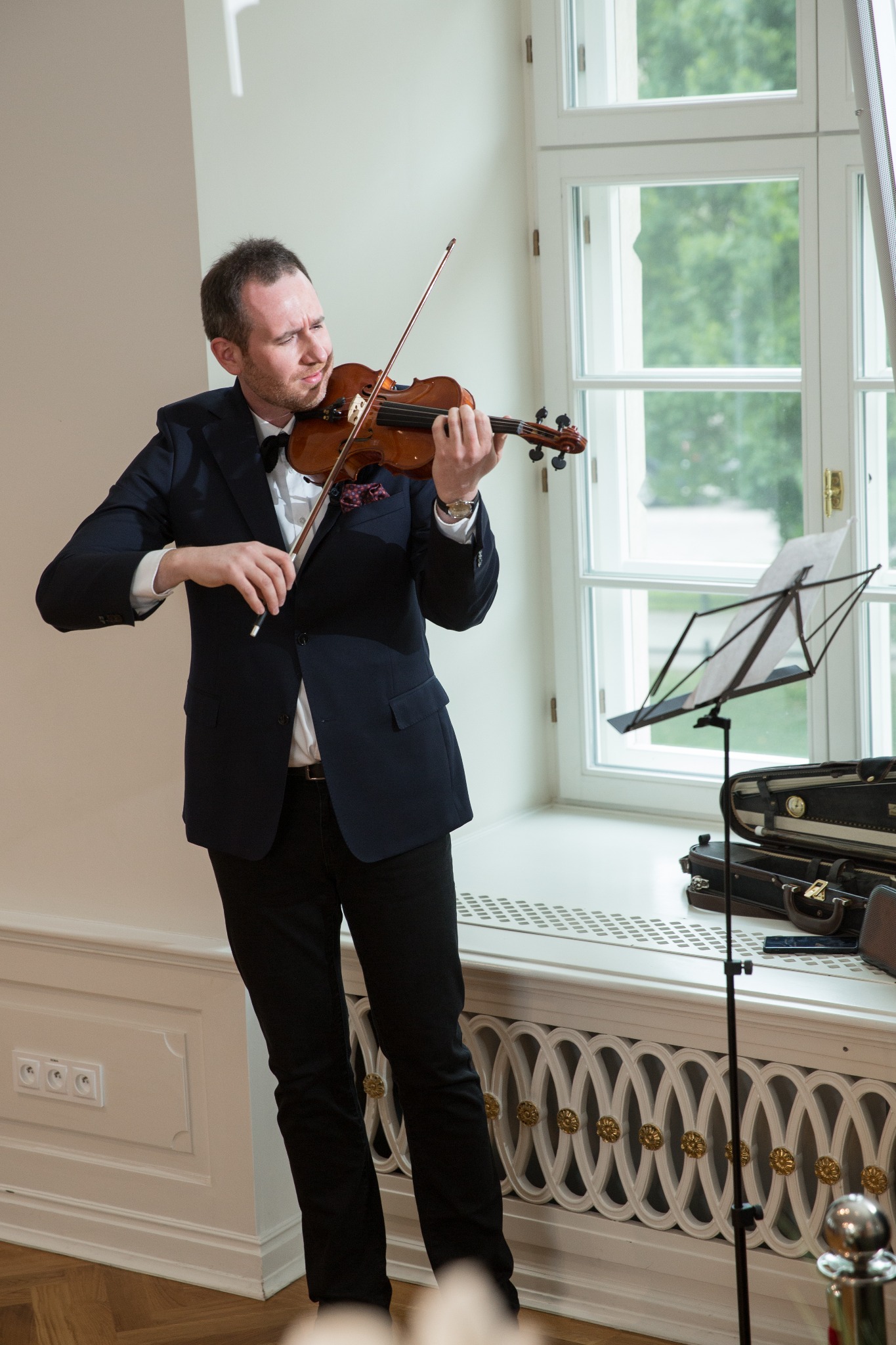 Skoncentrowany skrzypek w ciemnym garniturze z muszką gra na skrzypcach, nuty na statywie, otwarte etui na skrzypce w tle, jasne wnętrze z oknem.