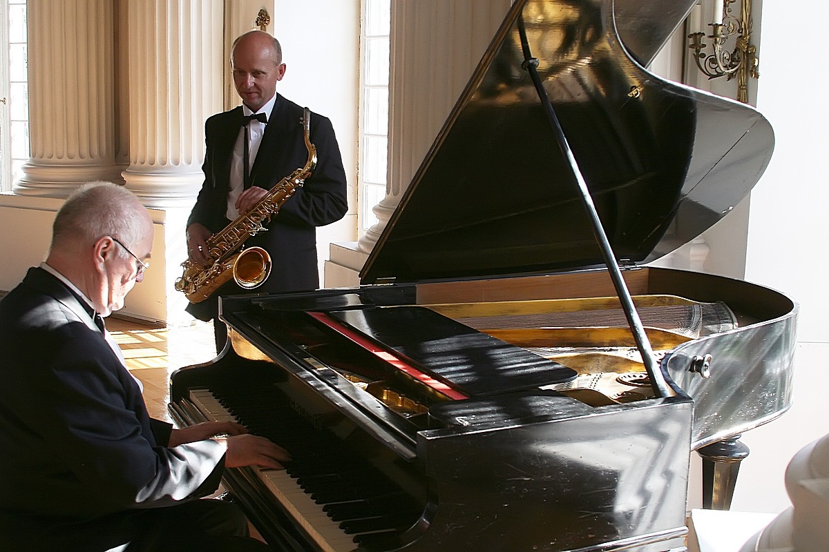 Duet muzyczny w eleganckim wnętrzu: pianista gra na czarnym fortepianie, a saksofonista w smokingu stoi obok, gotowy do występu, w tle kolumny i okna.