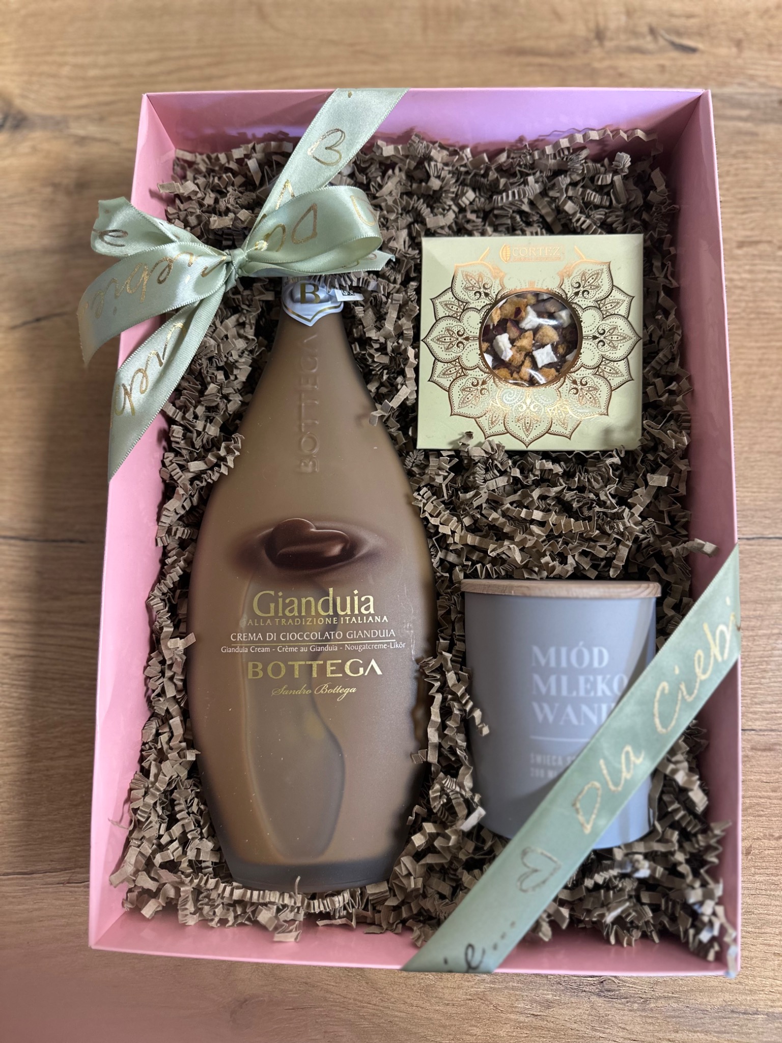Różowe pudełko prezentowe wypełnione brązowym wypełniaczem, zawierające butelkę kremu czekoladowego Gianduia, pudełko słodyczy i świecę sojową z napisem 'Miód Mleko Wanilia', przewiązane wstążką...