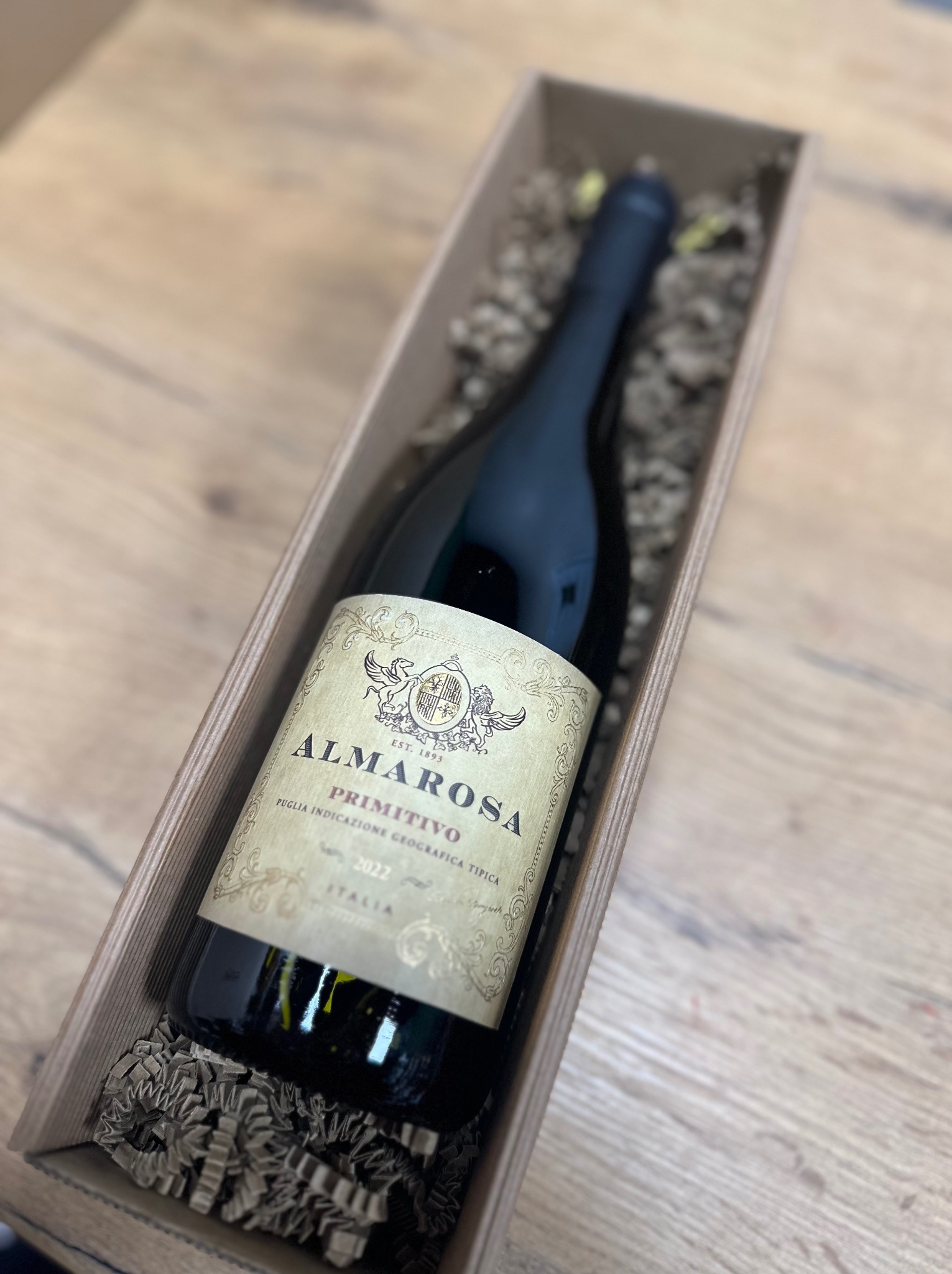 Butelka czerwonego wina Almarosa Primitivo 2022 w drewnianej skrzynce wypełnionej papierowym wypełniaczem na drewnianym stole.