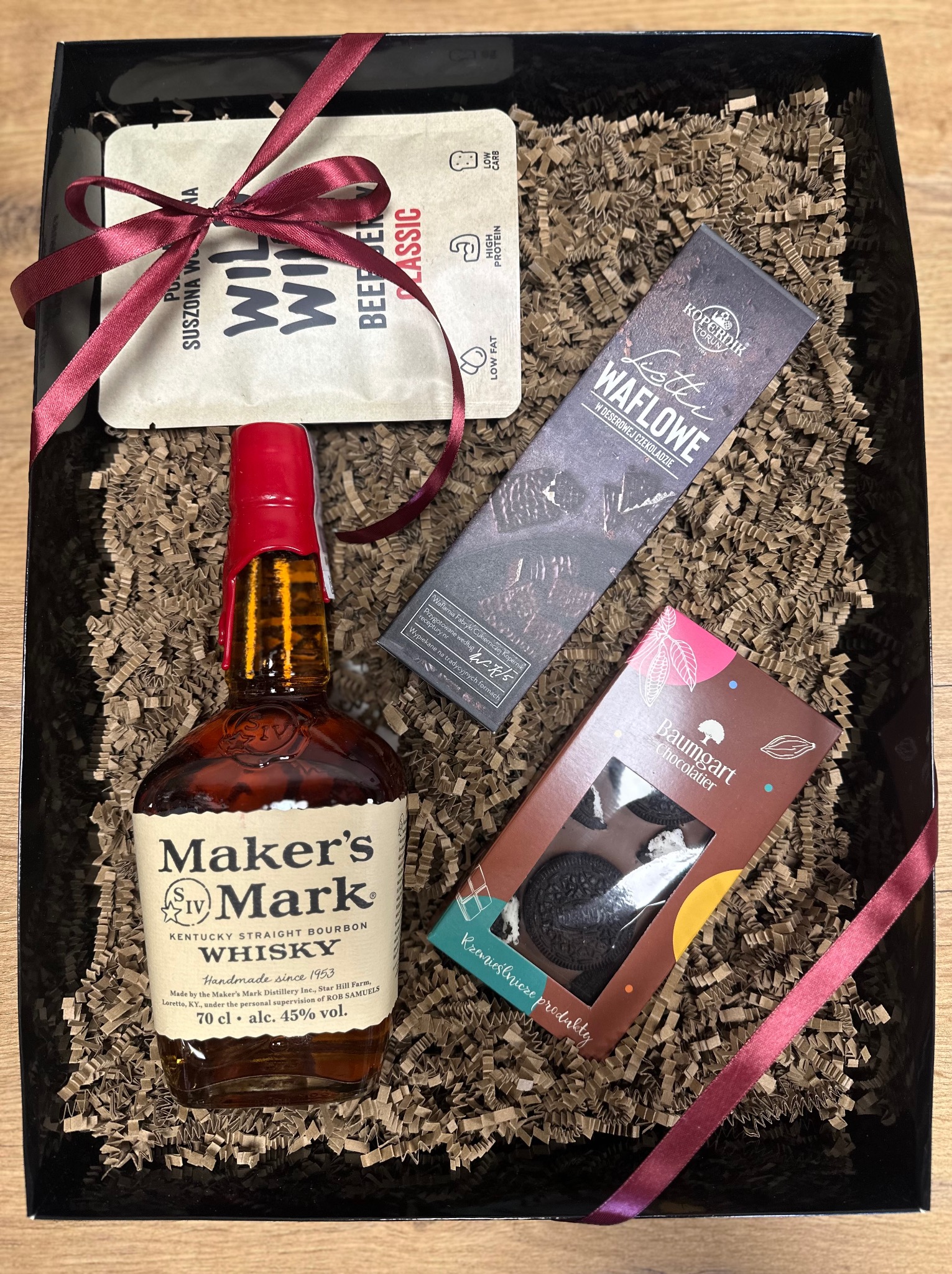 Czarny kosz prezentowy wypełniony brązowym wypełniaczem, zawierający butelkę whisky Maker's Mark z czerwonym korkiem, suszoną wołowinę, czekoladę Kopernik Listic Waflowe i czekoladki Baumgart...