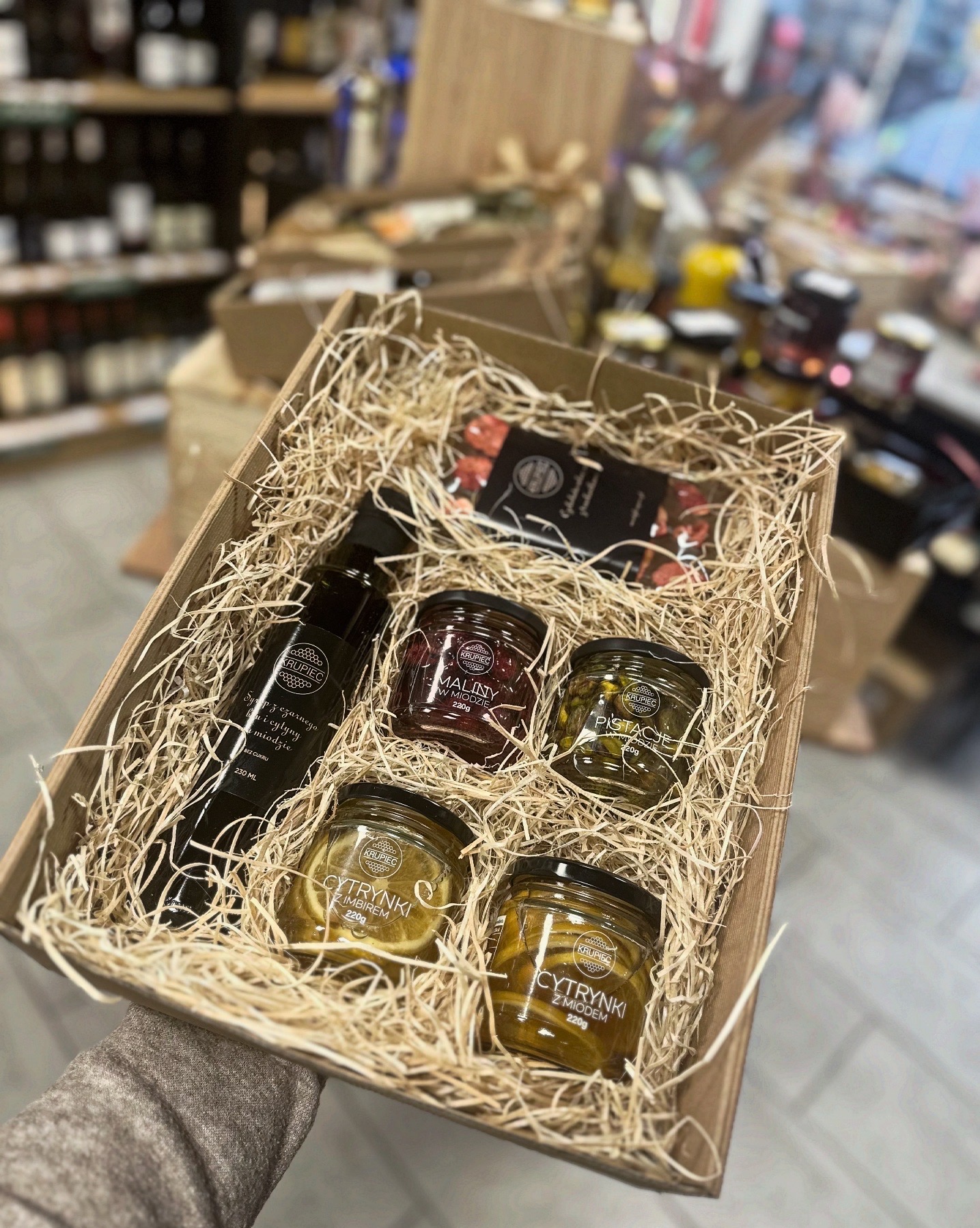Ręcznie wykonany, ekologiczny upominek: drewniany kosz wypełniony siankiem, zawierający butelkę syropu, słoiczki z cytrynami w miodzie, malinami w miodzie i pistacjami w miodzie oraz czekoladki.