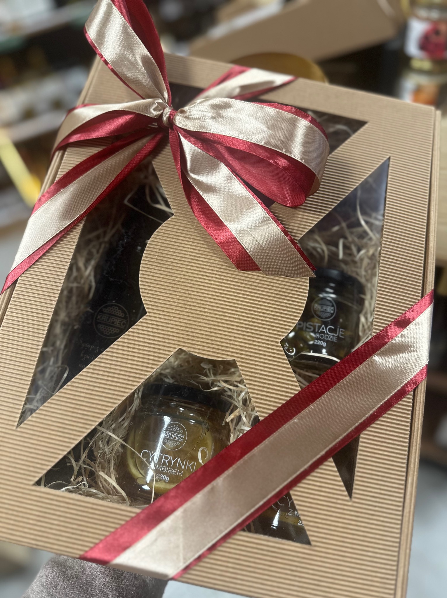 Kartonowe pudełko prezentowe z trzema słoiczkami miodu, przewiązane czerwoną i kremową wstążką, wypełnione siankiem.