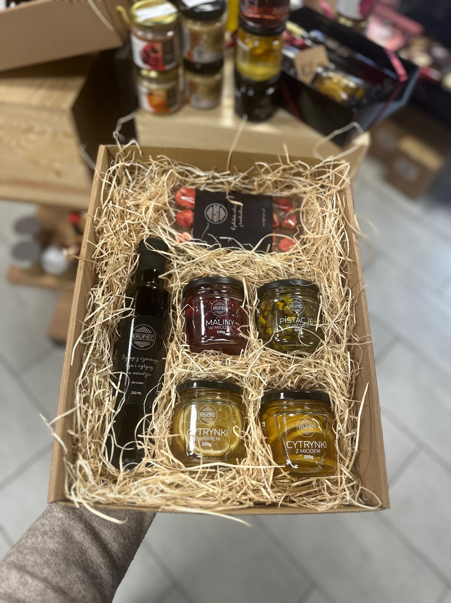 Ręcznie trzymany kartonowy box wypełniony siankiem, zawierający butelkę syropu i cztery słoiczki z przetworami owocowymi w miodzie: maliny, pistacje i cytryny z imbirem, całość tworzy gotowy zestaw...