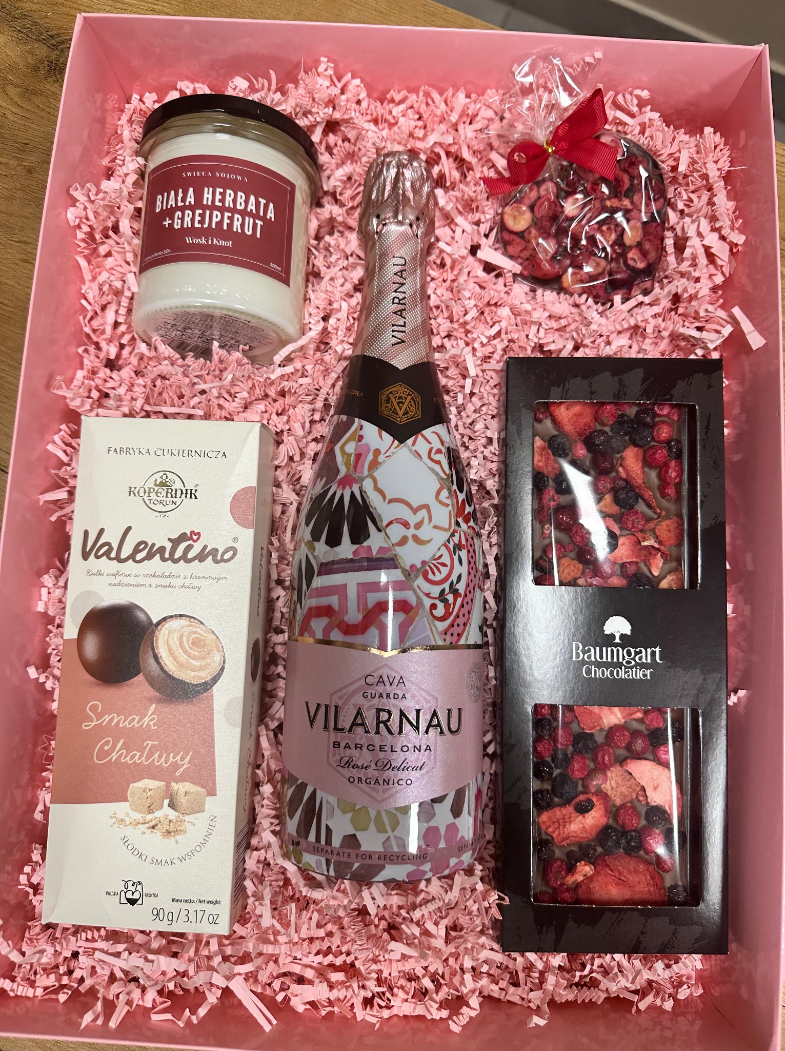 Różowy kosz prezentowy wypełniony dekoracyjnym papierem, zawierający butelkę różowego wina musującego Vilarnau, czekoladę Baumgart z owocami liofilizowanymi, praliny Valentino od Kopernika i świecę...