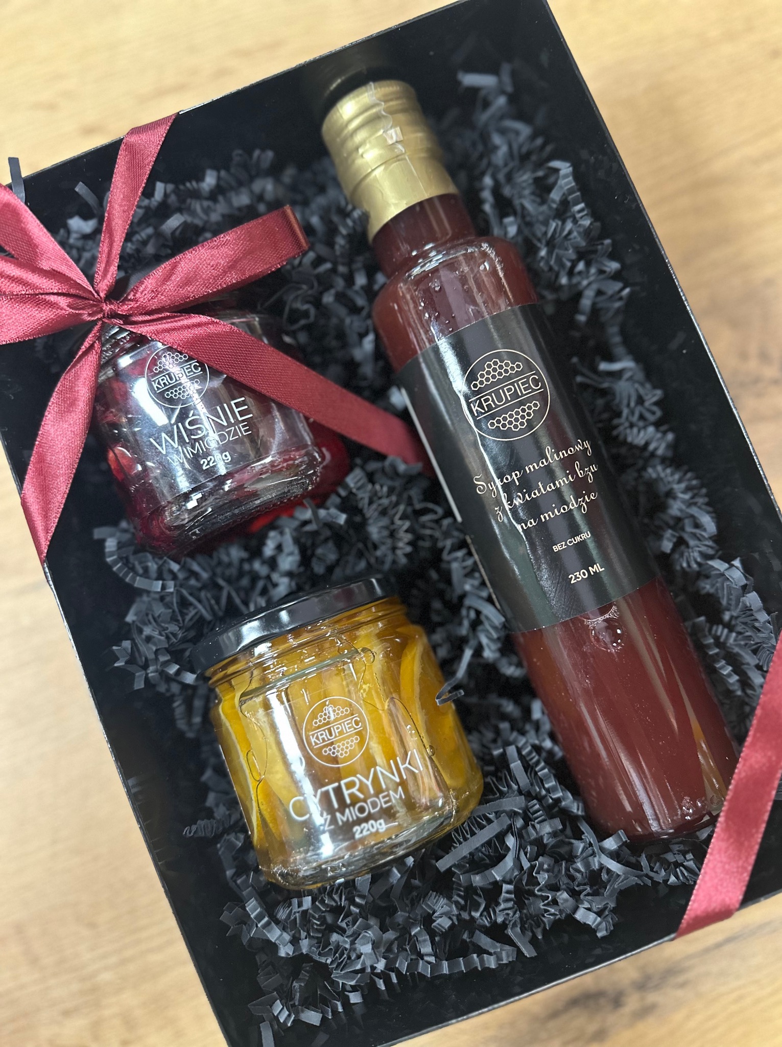 Elegancki czarny kosz prezentowy wypełniony czarnym siankiem, zawierający słoik z wiśniami w miodzie, słoik z cytrynkami w miodzie oraz butelkę syropu malinowego z kwiatami bzu na miodzie, wszystko...