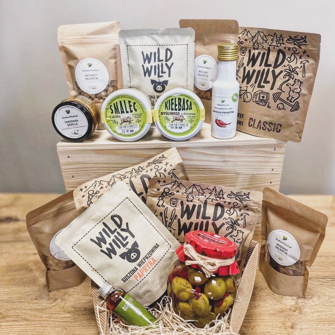 Zestaw produktów spożywczych marki Wild Willy w koszu prezentowym, zawierający smalec, kiełbasę, tapenadę, migdały, paprykę w słoiku i butelce, zapakowane w papierowe torebki i puszki, całość...
