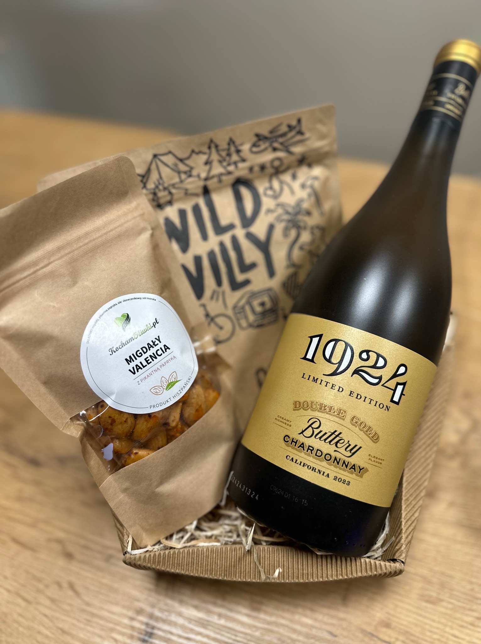 Elegancko zapakowany zestaw prezentowy: butelka białego wina Chardonnay 1924 Double Gold, migdały Valencia z pikantną papryką w brązowym papierowym opakowaniu oraz przekąski Wild Vily, umieszczone...