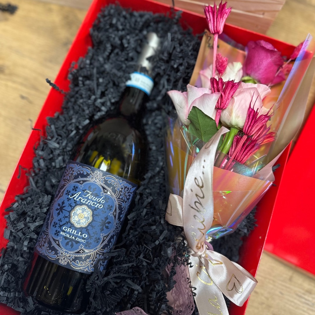 Czerwone pudełko prezentowe wypełnione czarnym wypełniaczem, zawierające butelkę wina Feudo Arancio Grillo Sicilia DOC oraz bukiecik różowych i białych róż, przewiązany wstążką z napisem 'Liebe...'