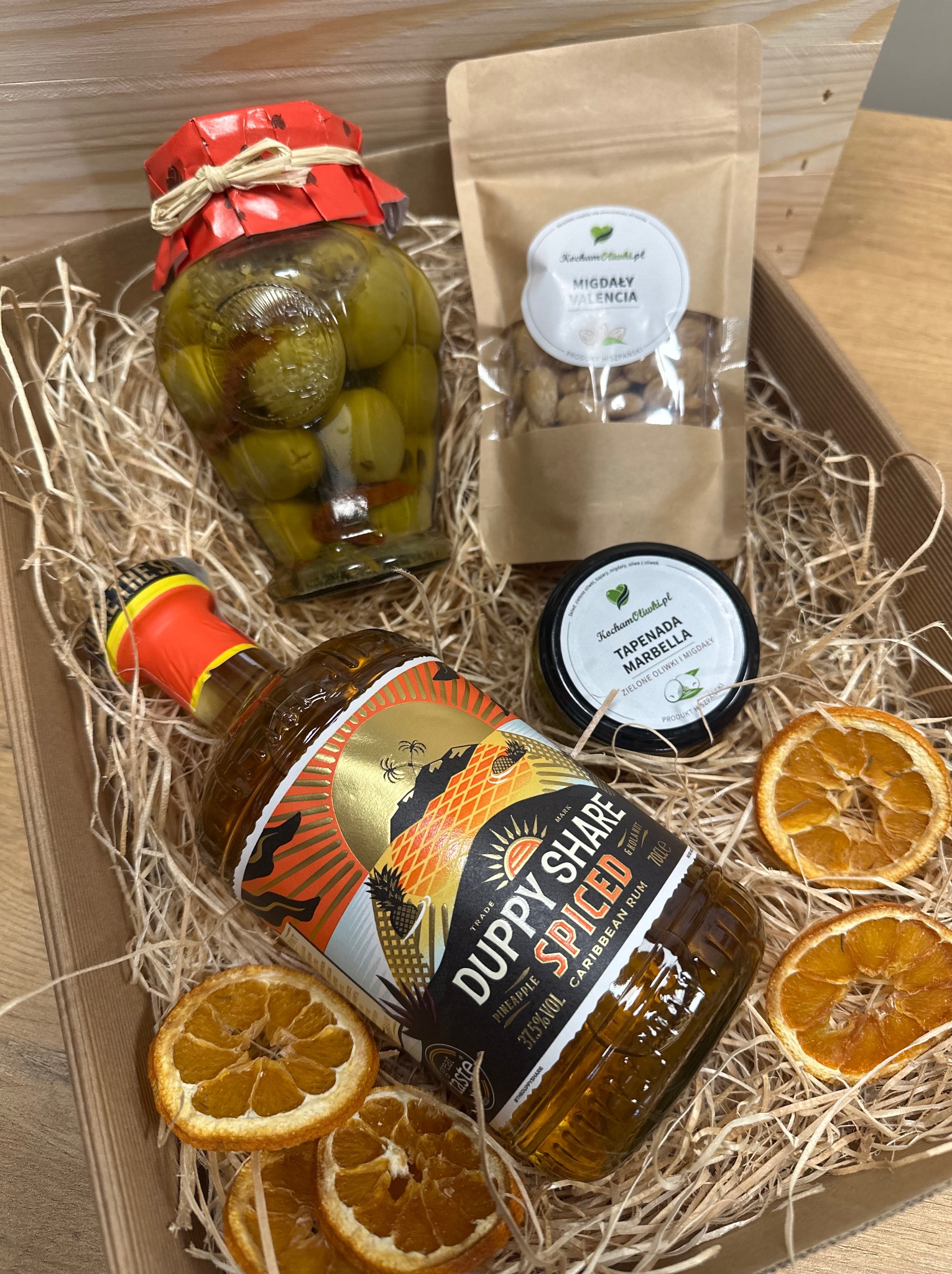 Zestaw prezentowy w kartonowym pudełku wypełnionym siankiem: butelka rumu Duppy Share Spiced, słoik zielonych oliwek, tapenada z oliwek i migdałów, migdały Valencia oraz suszone plastry pomarańczy.