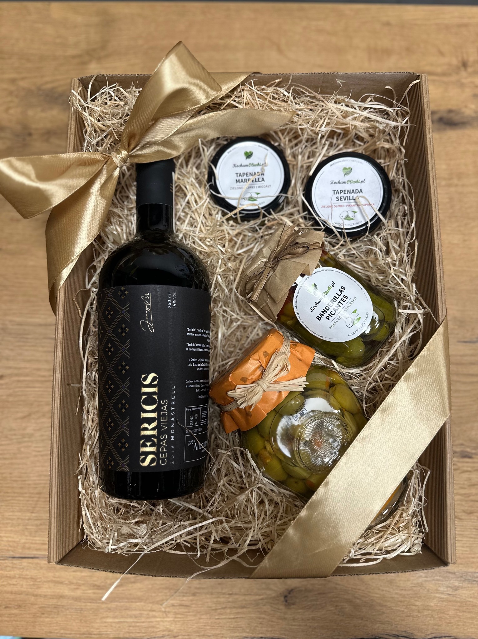 Elegancki kosz prezentowy z hiszpańskim winem Sericis, tapenadami z oliwek i koreczkami w słoikach, przewiązany złotą wstążką i wypełniony siankiem.