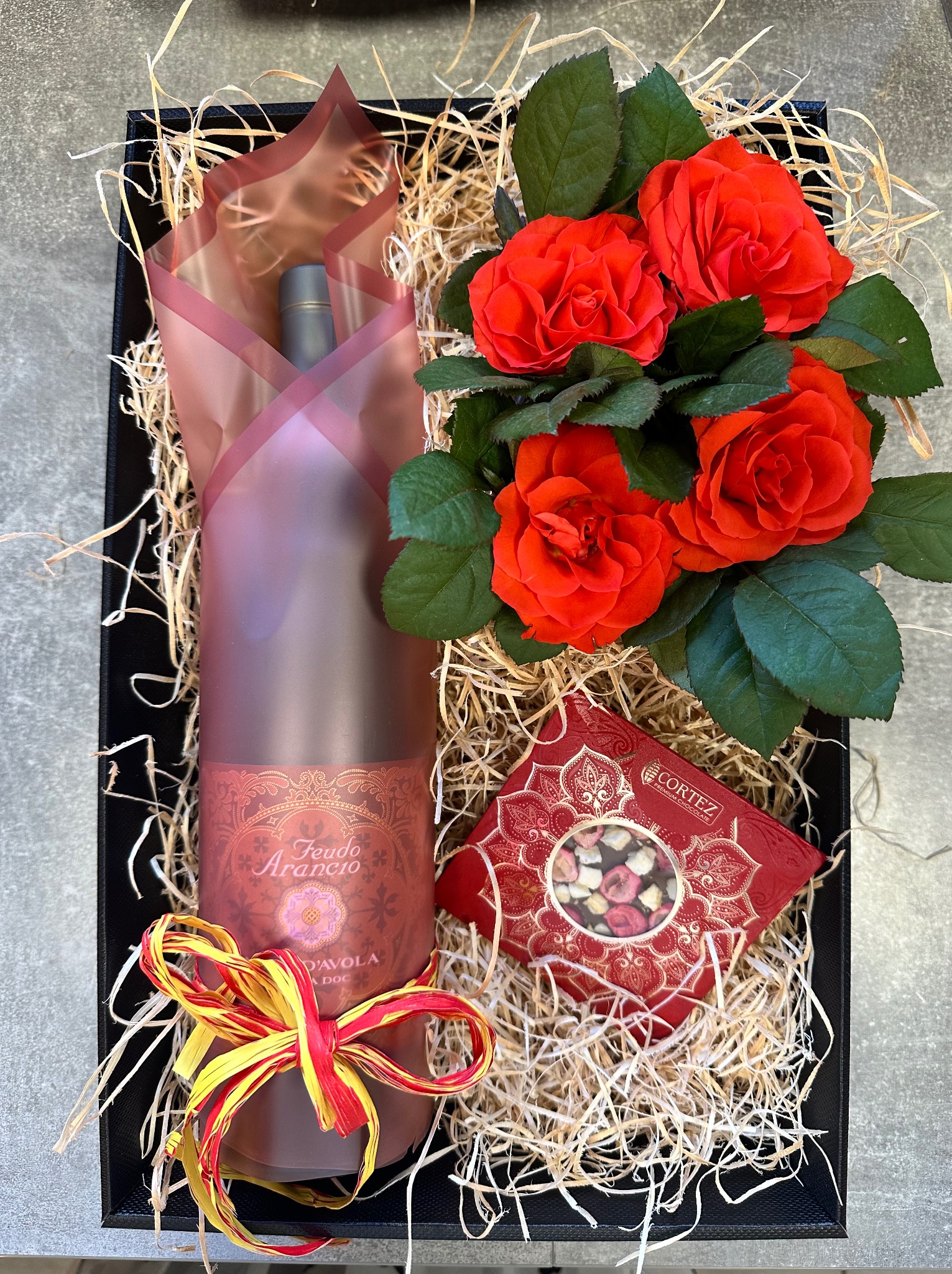 Elegancki czarny kosz prezentowy wypełniony siankiem, zawierający butelkę wina zawiniętą w ozdobny papier, pudełko czekoladek Cortez i bukiet intensywnie czerwonych róż.