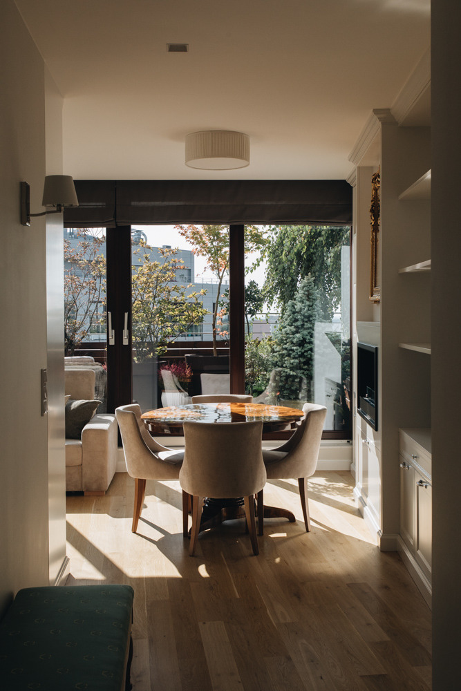 Jasne wnętrze jadalni z okrągłym stołem, krzesłami w kolorze beżowym i parkietem w promieniach słońca, widok na taras z roślinnością.