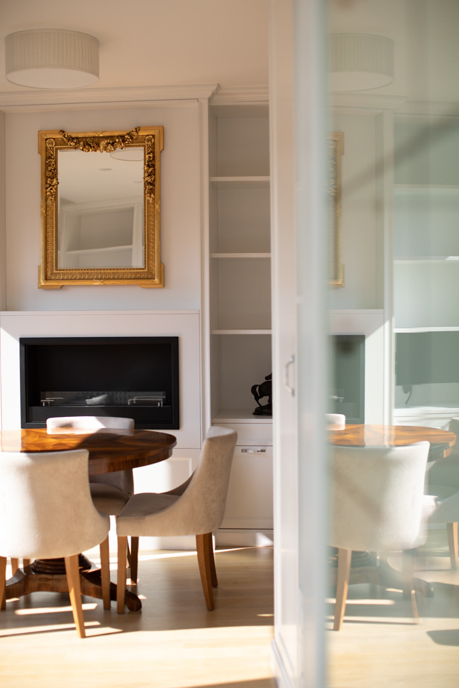Elegancka jadalnia z okrągłym stołem z ciemnego drewna, krzesłami w odcieniach beżu, wbudowanym regałem z półkami i kominkiem, lustro w złotej ramie na ścianie, widok przez przeszklone drzwi.