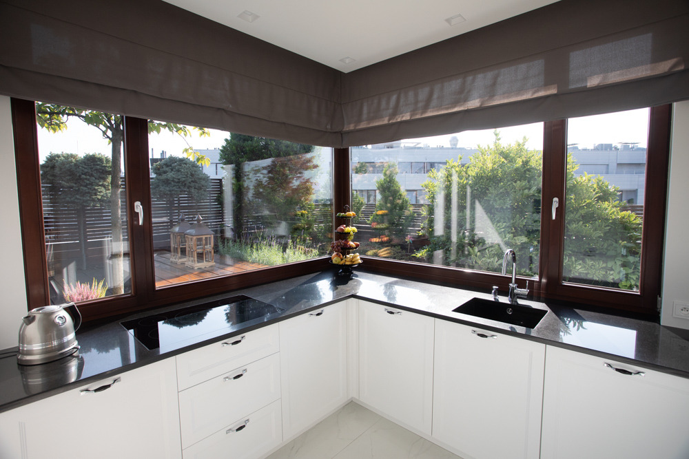 Nowoczesna kuchnia z białymi szafkami, czarnym blatem i dużym oknem narożnym z widokiem na taras i zieleń ogrodu.