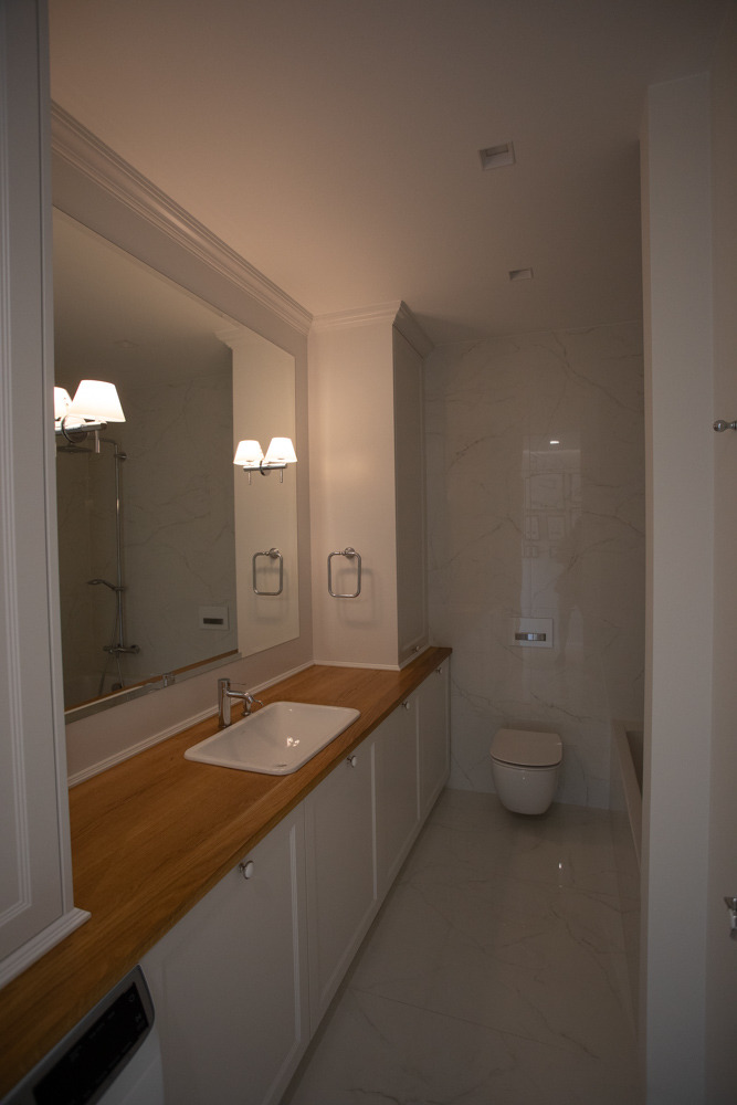 Nowoczesna łazienka z drewnianym blatem pod umywalką, białymi szafkami, marmurowymi płytkami na ścianach i podłodze oraz minimalistyczną toaletą.