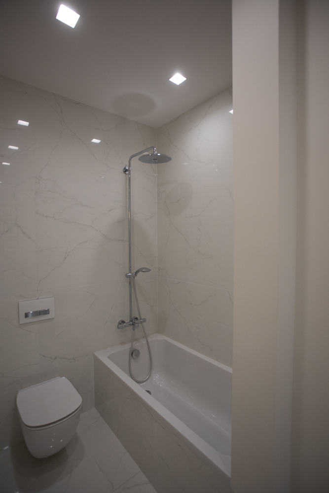 Nowoczesna łazienka z wanną obudowaną płytkami imitującymi marmur, wiszącą toaletą i chromowaną baterią prysznicową z deszczownicą.
