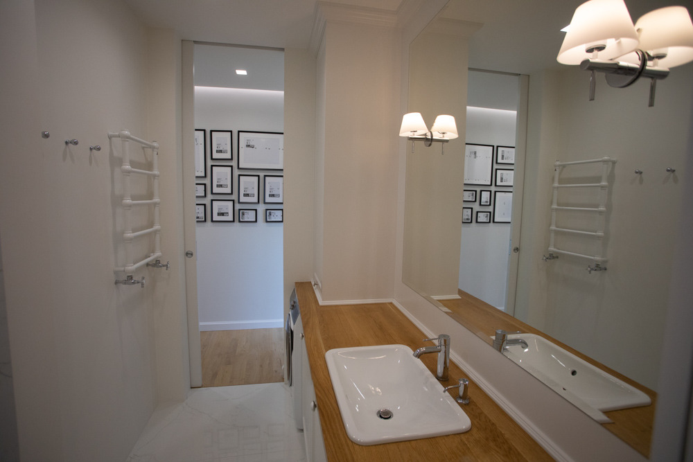 Nowoczesna łazienka w jasnych barwach z drewnianym blatem pod umywalką, widoczny grzejnik drabinkowy i galeria czarno-białych zdjęć w tle.